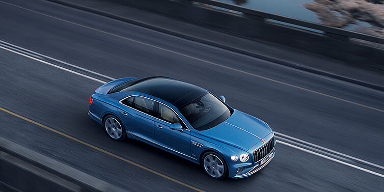 Bentley Porto New Bentley Flying Spur Azure sedan in Light windsor blue paint by Mulliner, with 22 inch Azure wheels seen from overhead driving dynamically on a country road