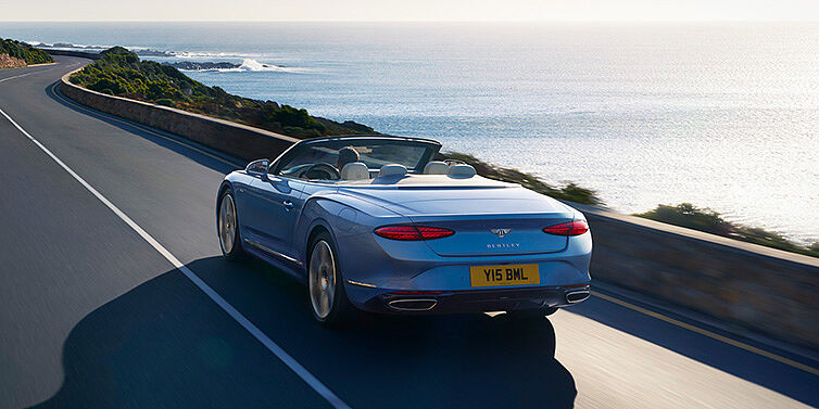 Bentley Porto New Bentley Continental GTC Azure convertible rear view in Light Windsor blue paint with 22 inch ten spoke wheels driving dynamically on an ocean road in the sunshine